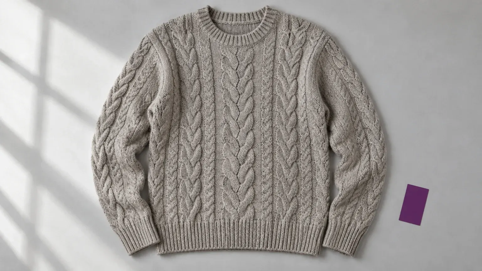 Overhead flat lay of a gray cable-knit sweater with both sleeves and hem visible, beside a purple card.