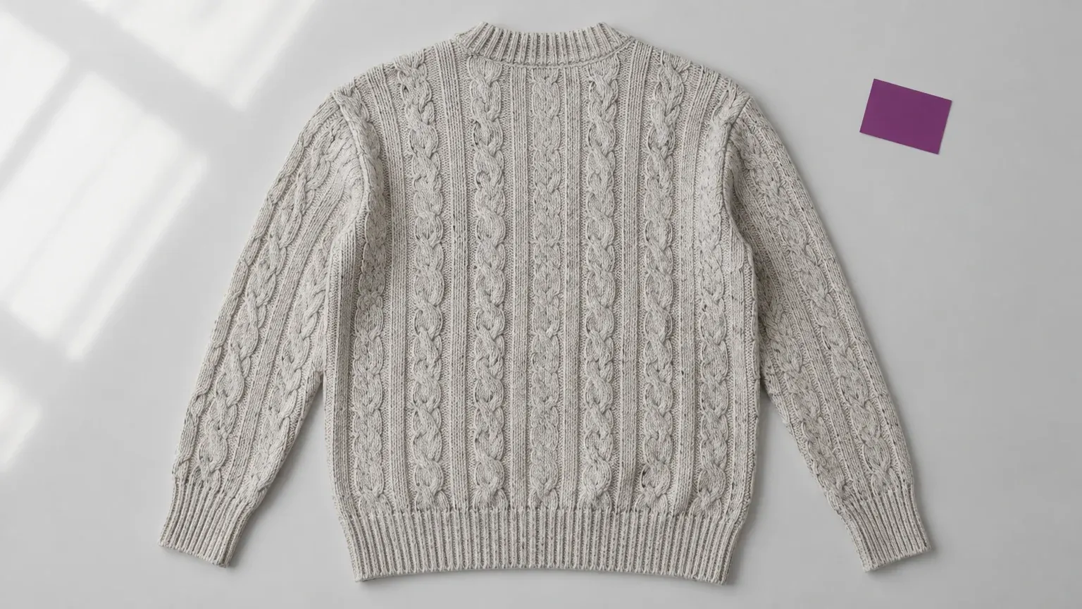 Overhead back view of a gray cable-knit sweater laid flat, with full sleeves and hem visible beside a purple card.