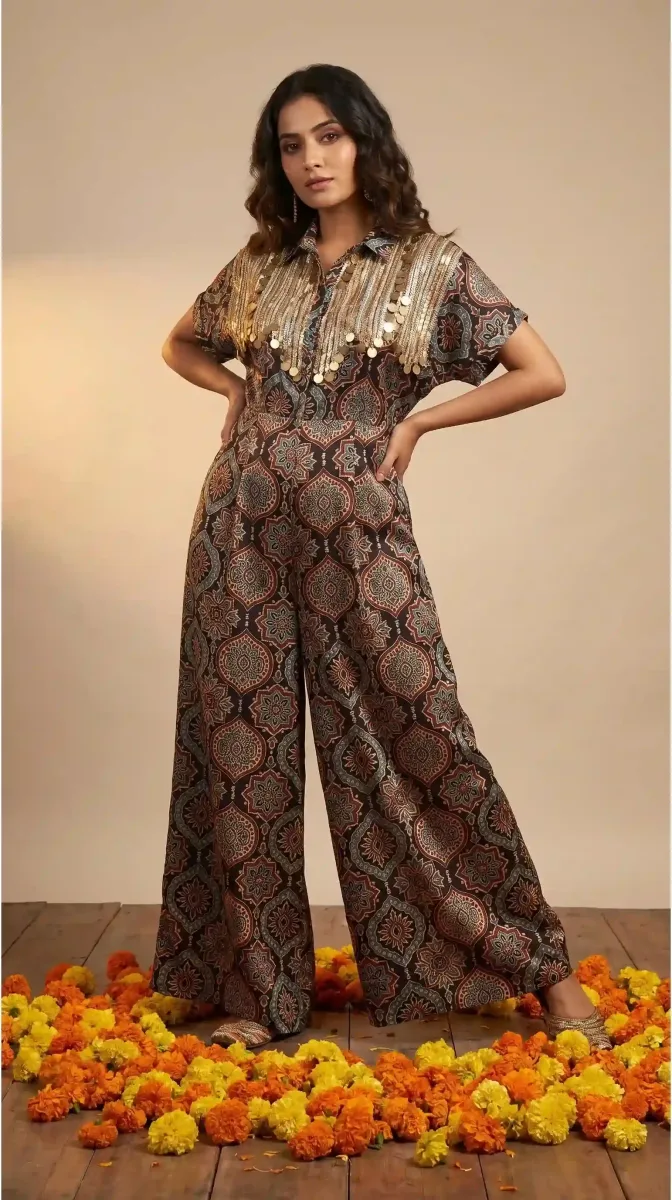 Model wearing a geometric-print wide-leg jumpsuit with gold-toned shoulder embellishments, standing among marigolds.